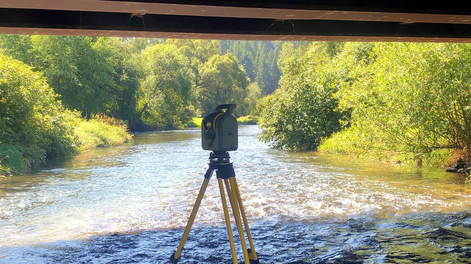 survey theodolite set in a stream under a bridge with scenic background